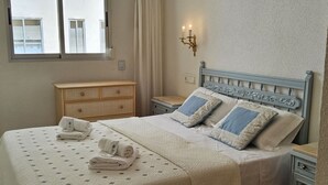 A neatly made bed with a quilt, pillows, and folded towels. There is a wooden dresser, a nightstand, and a wall-mounted light fixture. - Beautiful apartment for 4 guests with WIFI, A/C, TV, terrace and panoramic view (Altea)