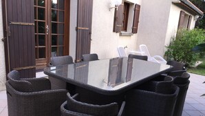 Een glazen eettafel omringd door rieten stoelen, op een betegeld terras met een gebouw op de achtergrond.