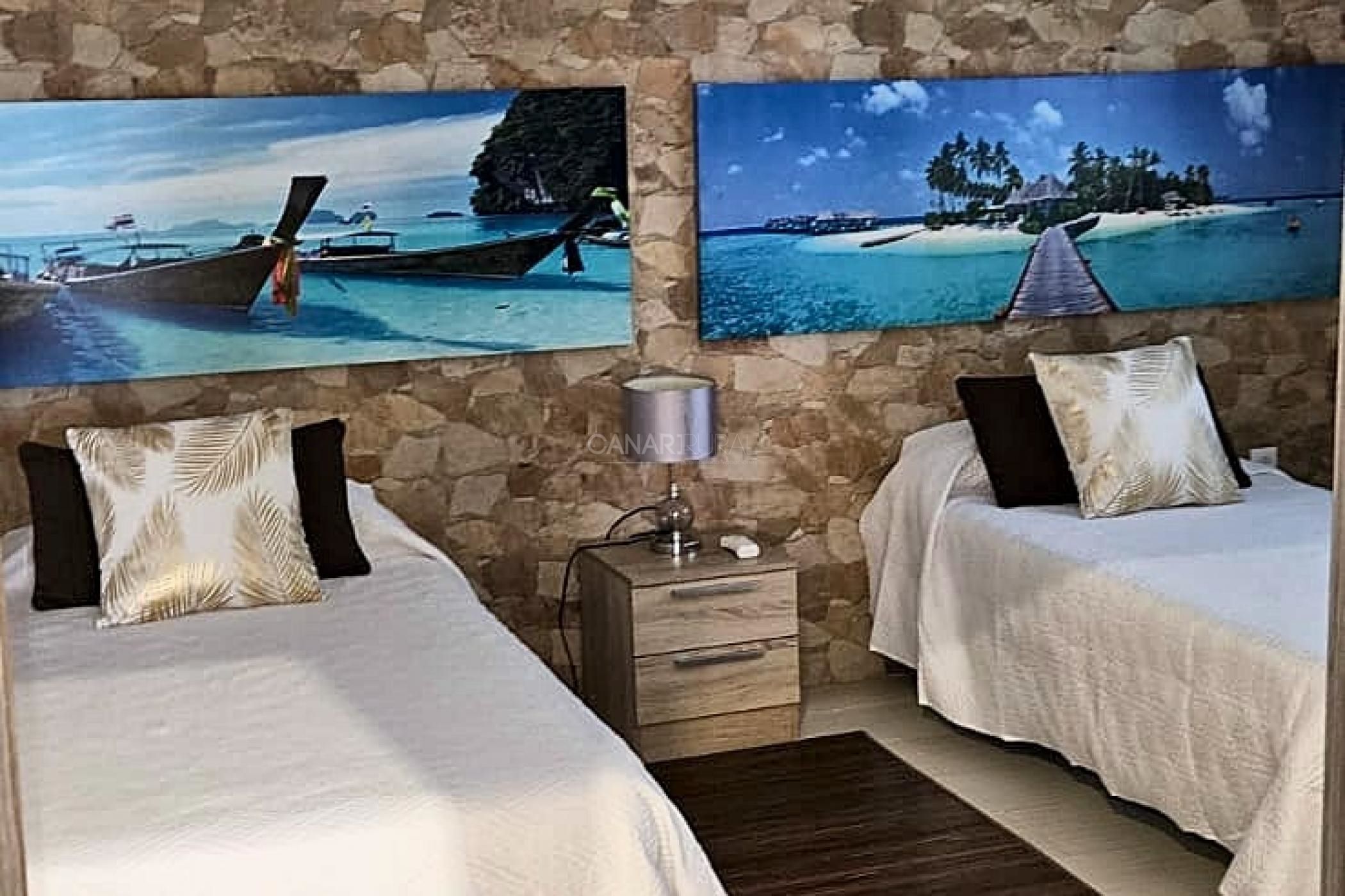 A hotel room with two beds, a nightstand, and a lamp. There are two paintings of tropical scenes on the wall.