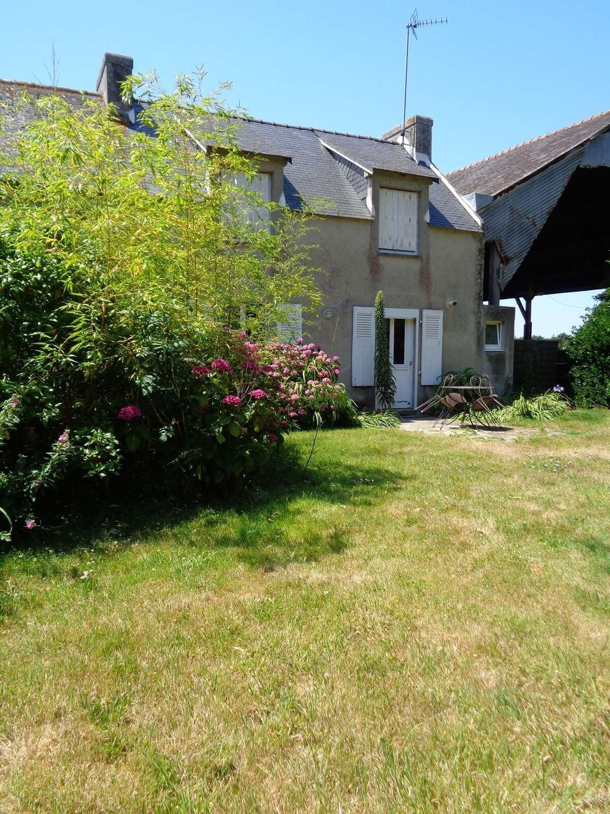 Une maison à deux étages dotée d’un porche couvert, d’une cheminée et d’un jardin agrémenté d’arbustes en fleurs.