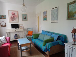 A cozy living room with a sofa, a small table, a TV on a desk, and various framed pictures on the walls.