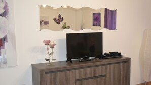 A modern living room with a flat-screen TV on a wooden cabinet, a decorative mirror with butterfly artwork, and a vase with flowers. - 1 bedroom apartment Las Caletillas (Las Caletillas)