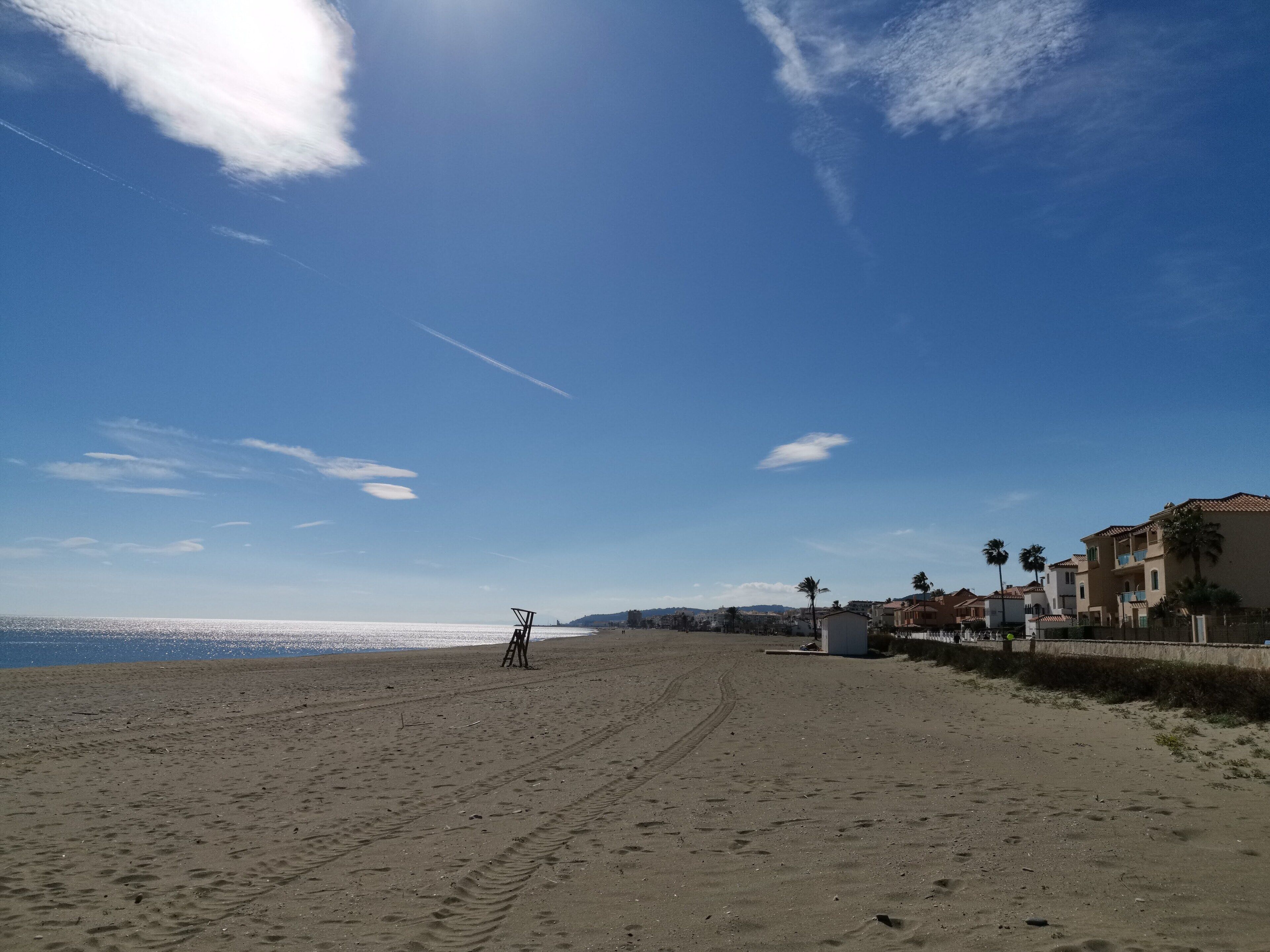 Een breed zandstrand met voetafdrukken, een heldere lucht en gebouwen op de achtergrond.