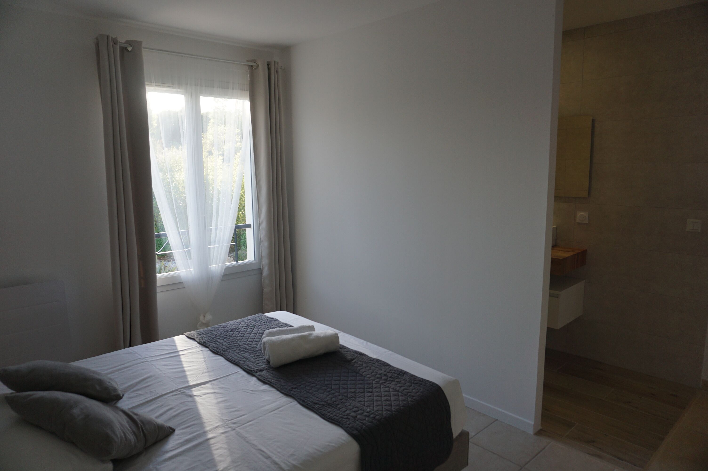 Un dormitorio con cama, ventana con cortinas y baño con espejo y lavamanos.