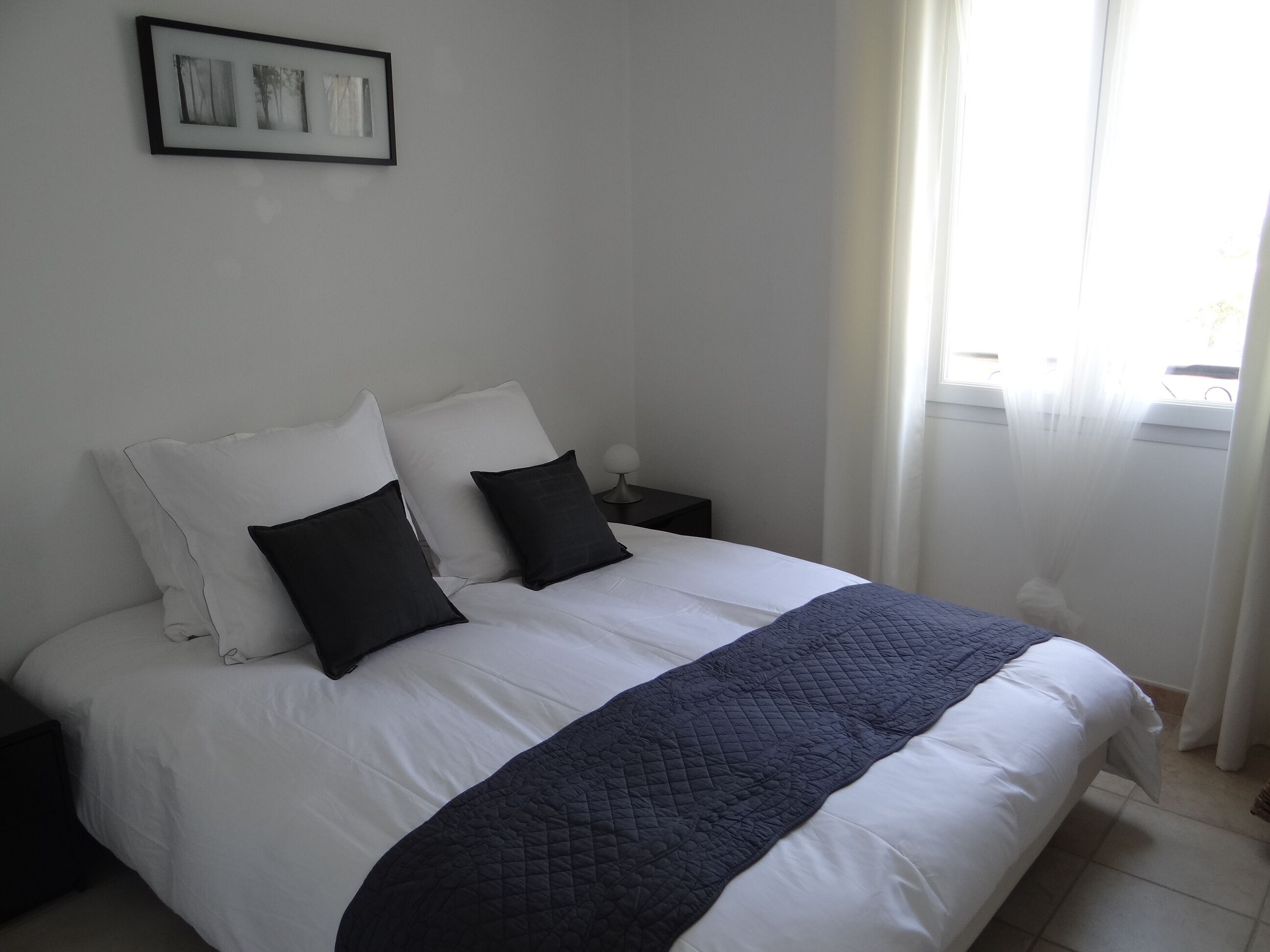 A neatly made bed with white and dark pillows, a dark quilted blanket, and a framed picture on the wall.