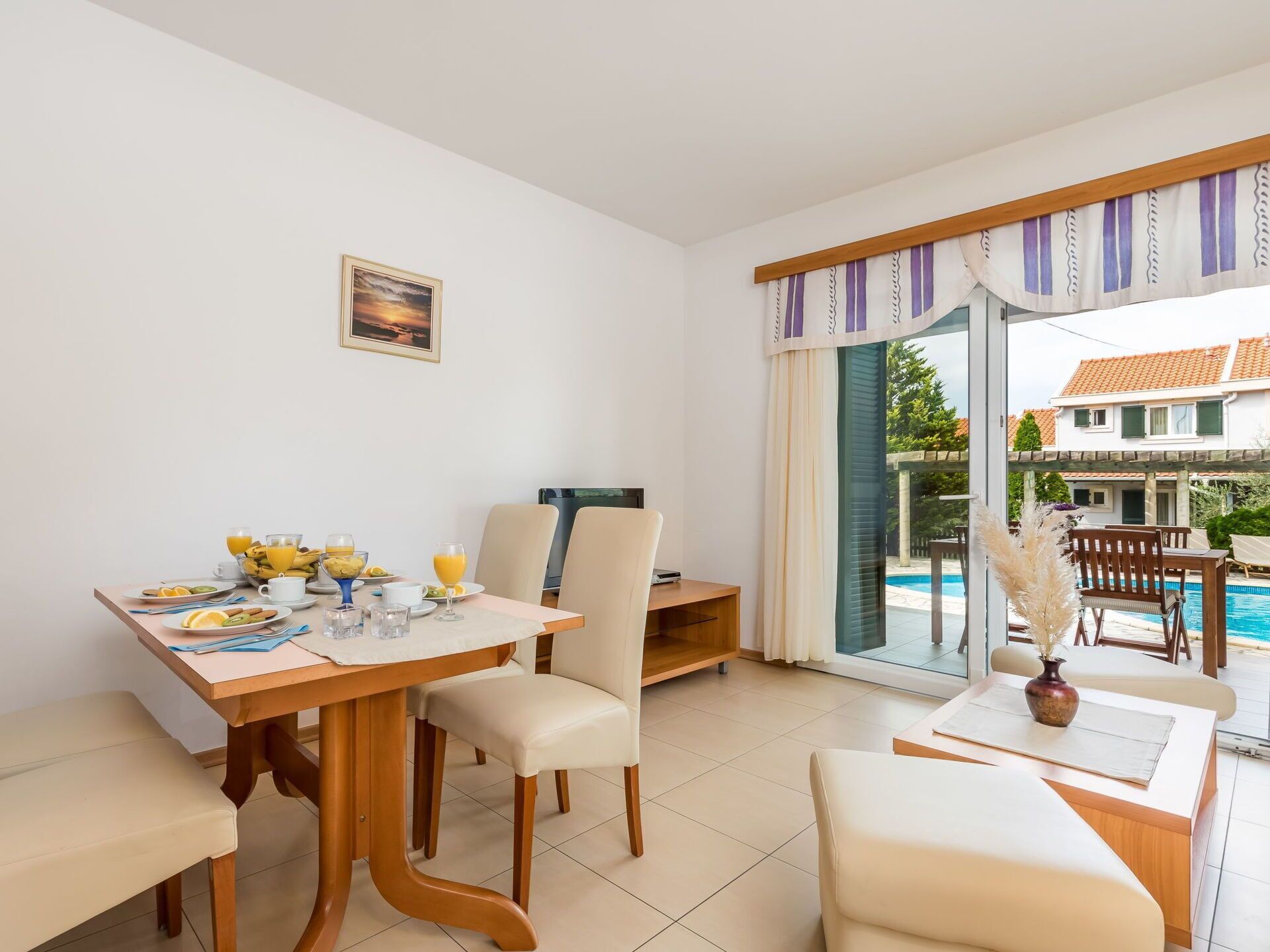 A dining area with a table set for breakfast, a TV, and a view of a swimming pool and house.