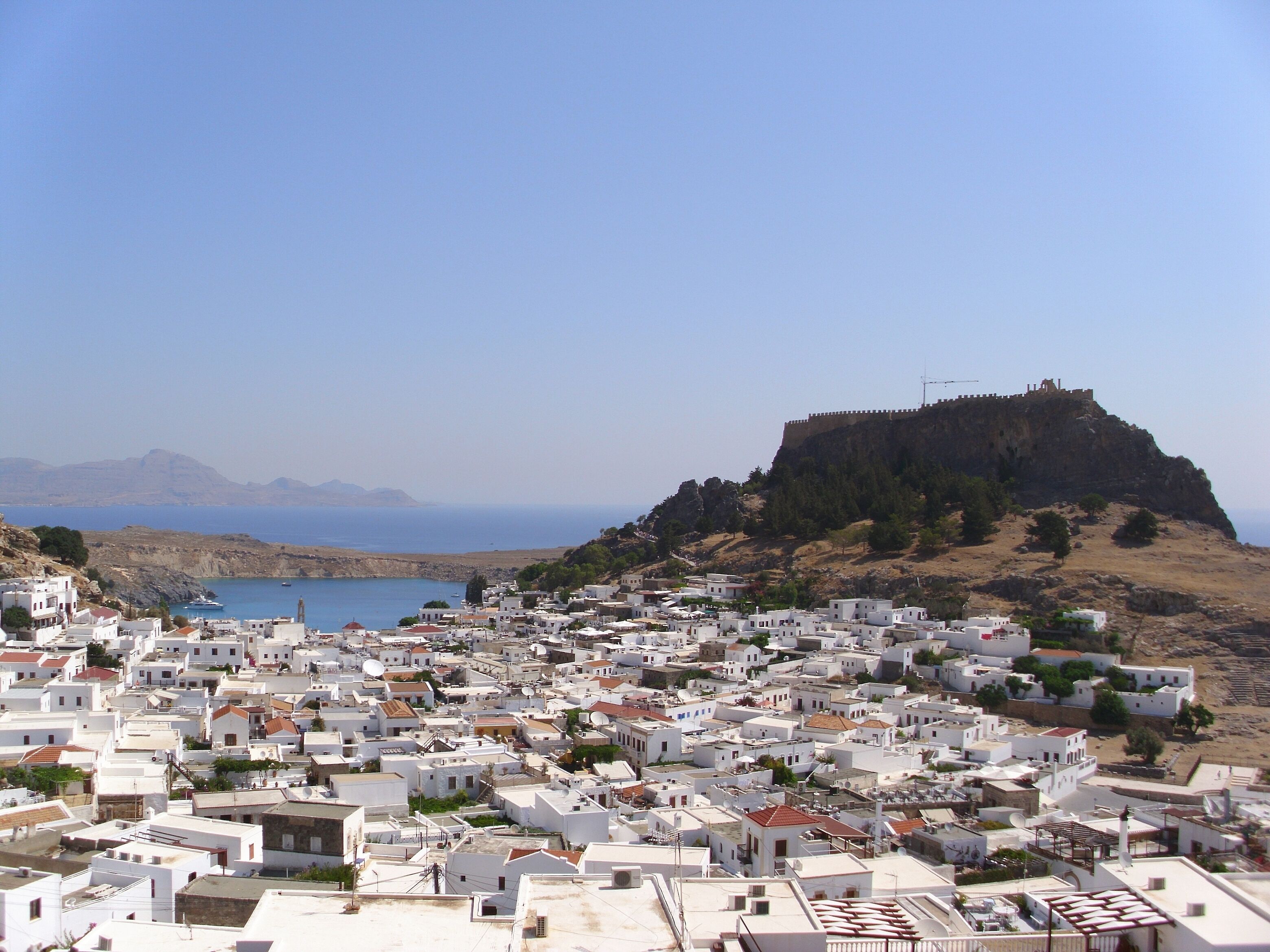 Un poble costaner amb edificis blancs, una fortalesa al cim d'un turó i mar blava i transparent.