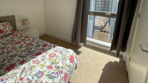 A bedroom with a bed, a nightstand, a window with curtains, and a view of buildings outside. - LUXURY SERVICED APARTMENTS***** (Croydon)