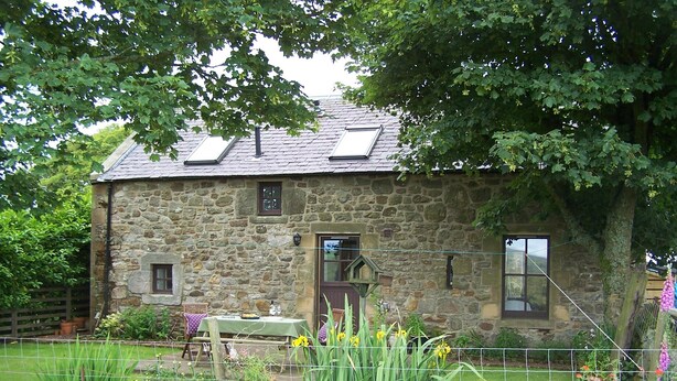 Une maison en pierre avec un toit en pente, un petit porche et une table avec une chaise à l’extérieur.