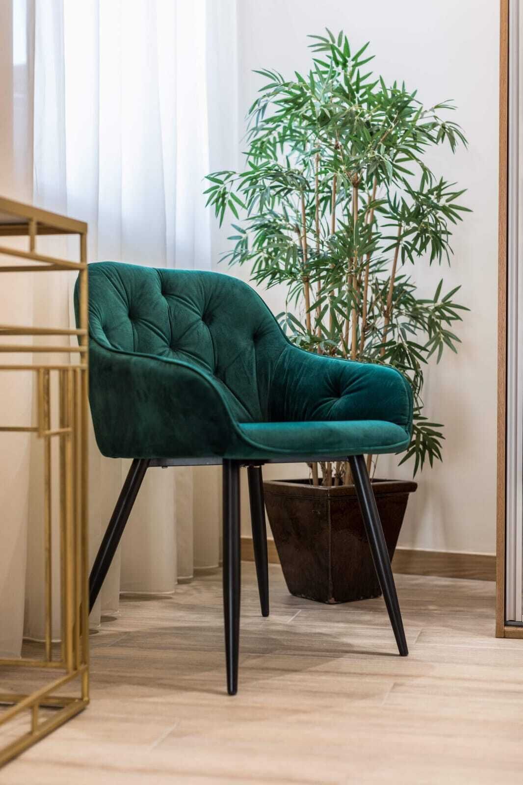 A teal velvet armchair with black legs, positioned next to a large bamboo plant in a dark pot, in a room with light wooden flooring and a gold-framed side table.