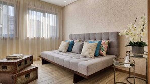 A modern bedroom with a large bed, a wooden chest of drawers, a small round table, and a potted plant. - Invicta Soul Joy (Porto)