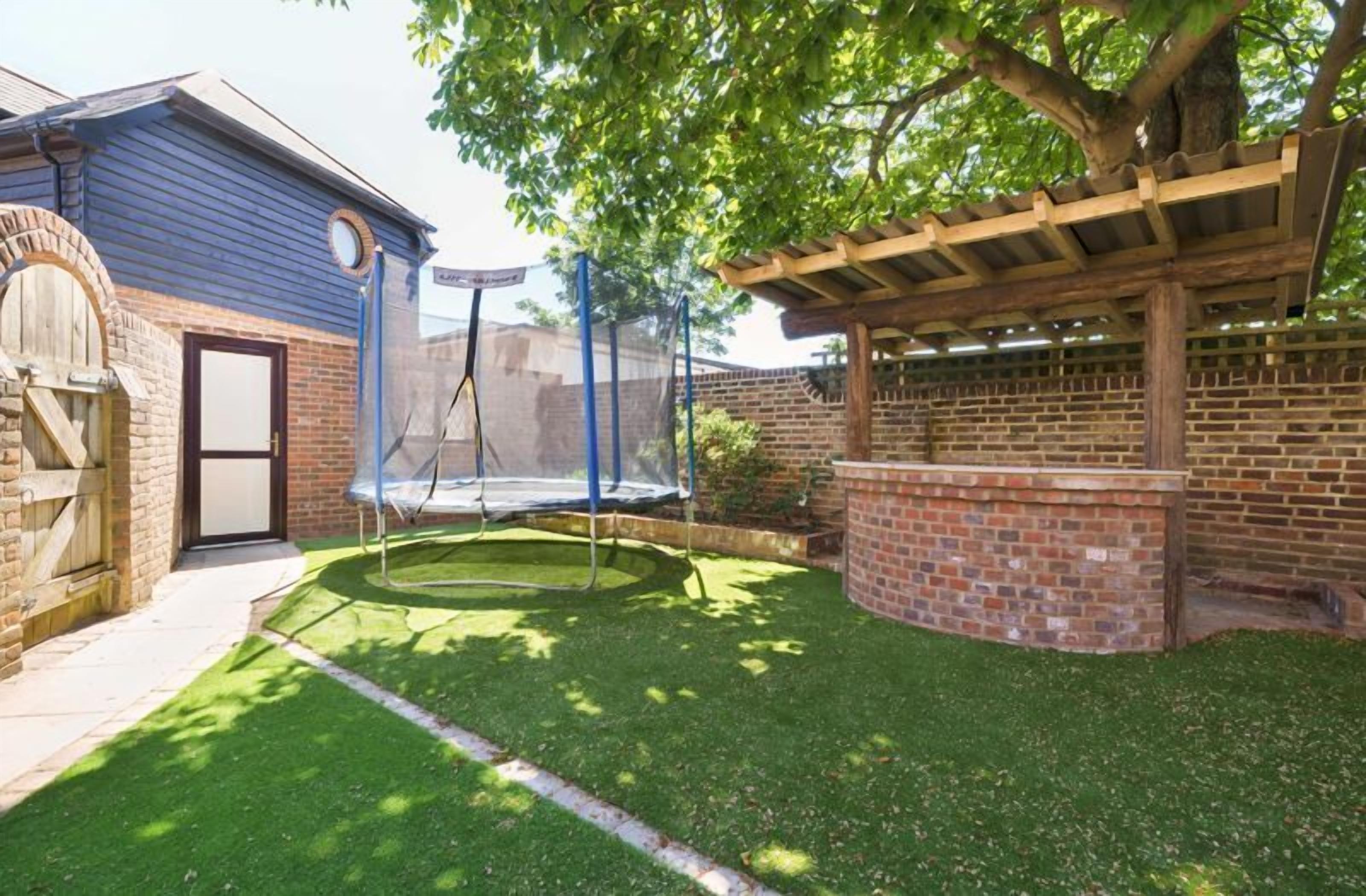 Patio trasero con un trampolín, un muro de ladrillo y una pérgola de madera.