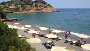 A beach with umbrellas and sun loungers, a rocky hill in the background, and calm sea.