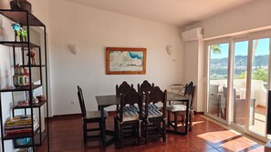 A dining area with a table and chairs, a bookshelf, a painting, and a view through a glass door.