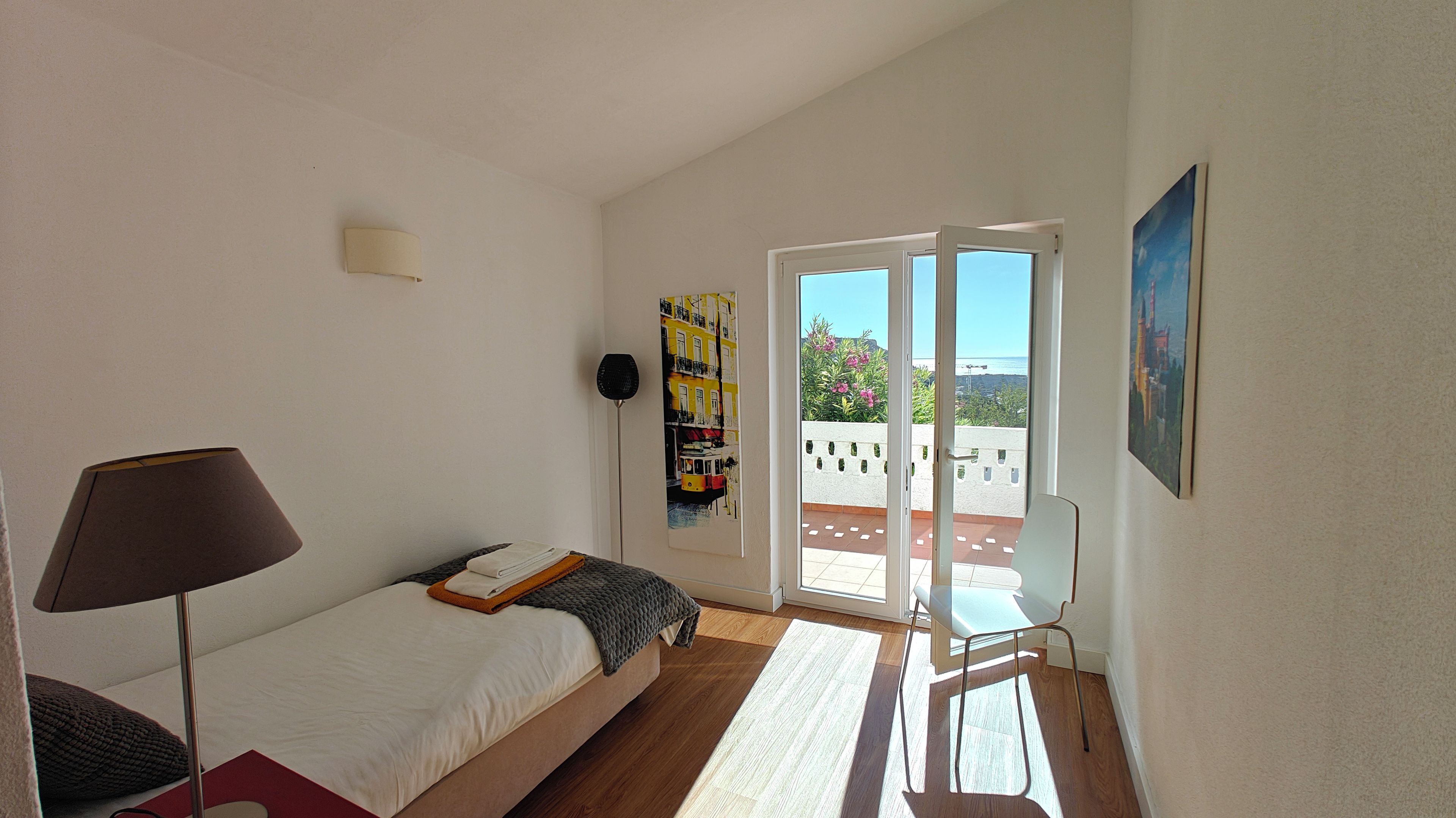 A bedroom with a bed, a lamp, a painting, and a view of the outdoors.