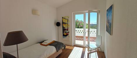 A bedroom with a bed, a lamp, a painting, and a view of the outdoors.