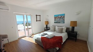 A bedroom with a bed, two bedside tables, a painting, and a view of the sea through a sliding door. - Villa Bela Vista T3 +1 (Lagos)