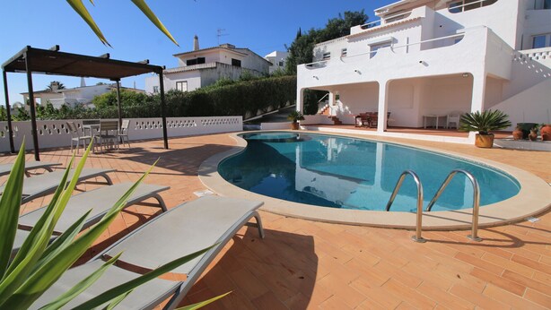 A swimming pool with lounge chairs, a covered seating area, and a white building with a tiled roof.