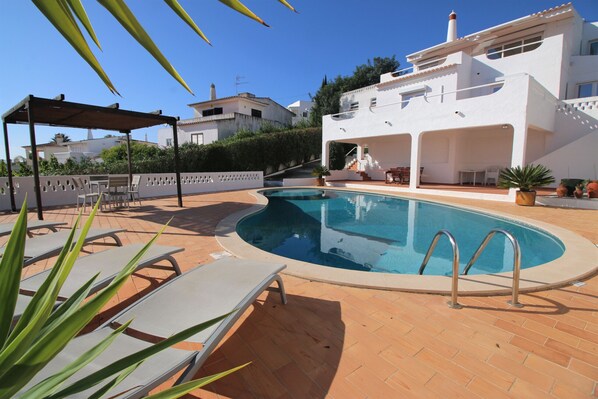 Une piscine avec des chaises longues, un espace de détente couvert et un bâtiment blanc doté d’un toit en tuiles.