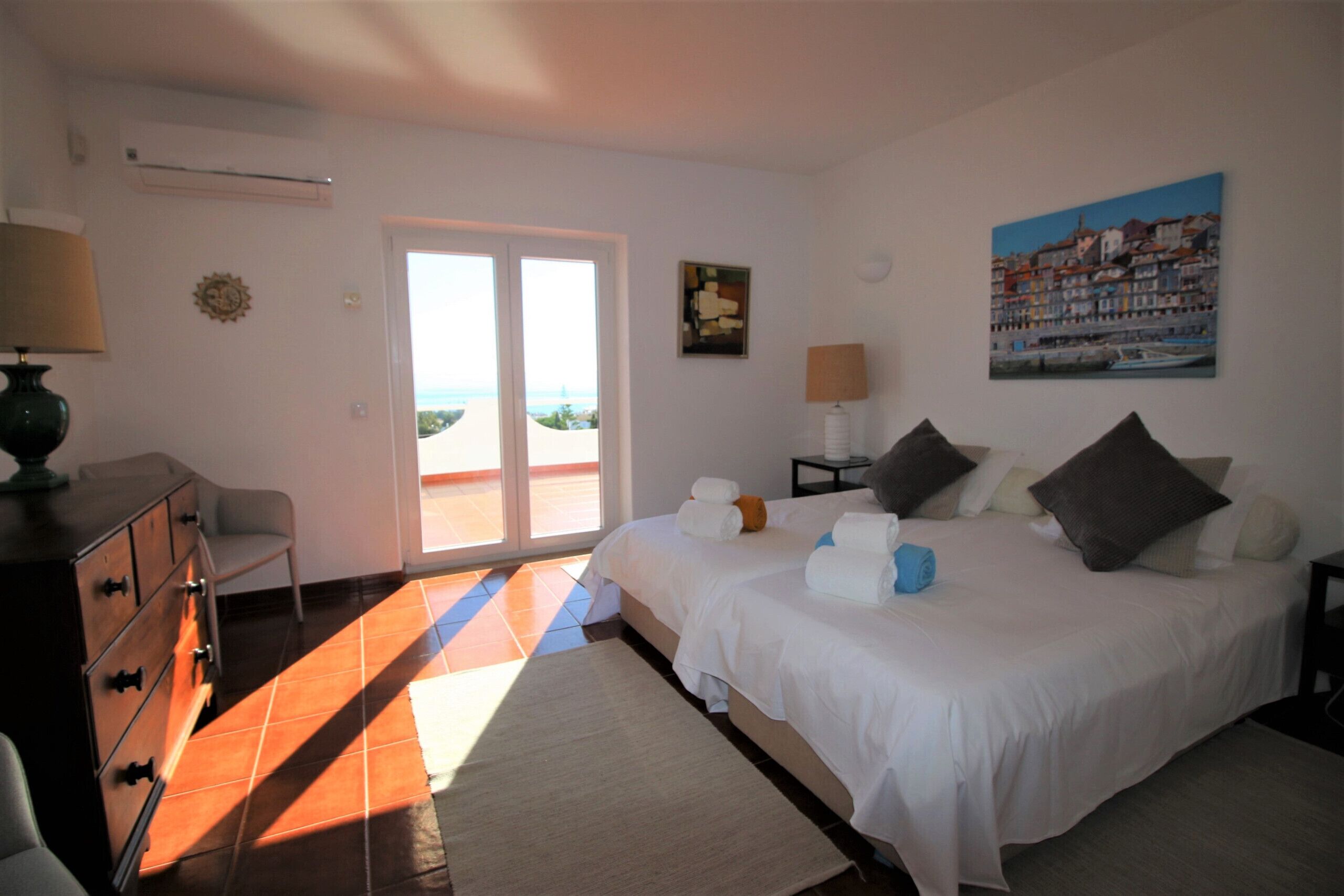 Une chambre à coucher comprenant un lit, une commode, une table de chevet, une lampe, un tableau et une vue sur l’extérieur, visible à travers une porte vitrée.
