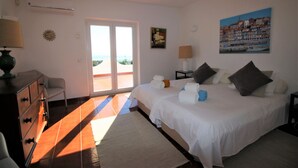 Ein Schlafzimmer mit Bett, Kommode, Nachttisch, Lampe, einem Gemälde und Blick durch eine Glastür.
