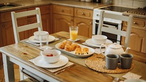 Una mesa de desayuno servida con cruasanes, jugo y tazas de té en una cocina con alacenas de madera y salpicadero de azulejos.