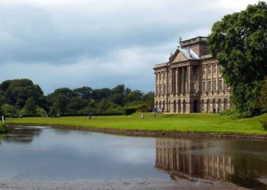 Un gran edificio de estilo clásico con fachada simétrica y ventanas arqueadas, rodeado de un césped cuidado y una masa de agua tranquila que refleja la estructura.