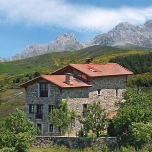 Un bâtiment en pierre avec un toit rouge, entouré de verdure et avec des montagnes en arrière-plan.