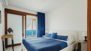 Ein Hotelzimmer mit einem großen Bett, einem Nachttisch, einem kleinen Tisch und Blick auf das Meer.