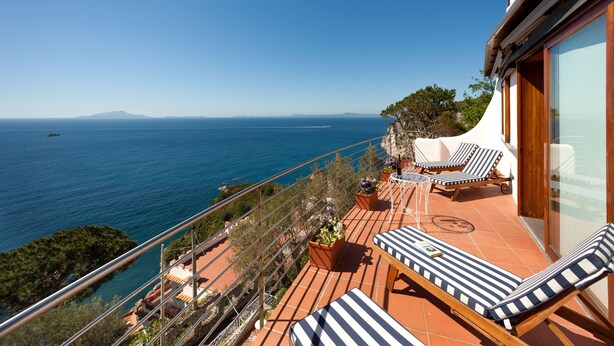 Ein Balkon mit Liegestühlen und Topfpflanzen mit Blick auf ein klares, blaues Meer.