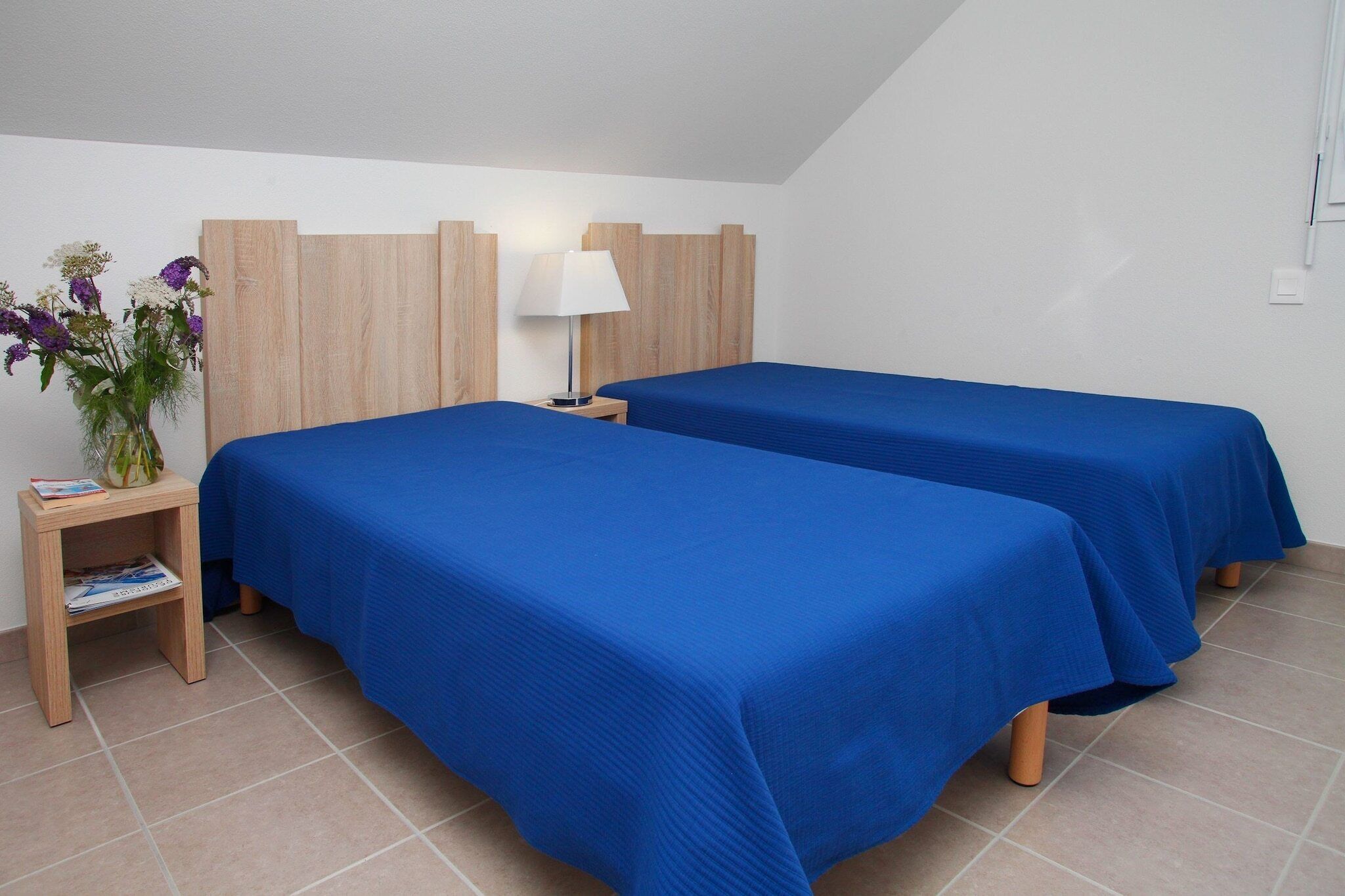A hotel room with two beds, a nightstand with a lamp, and a small table with a vase of flowers.