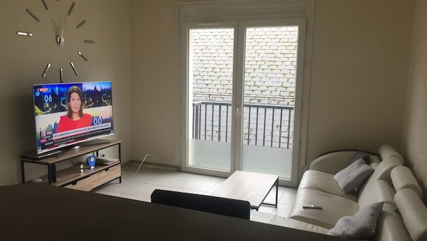 Un salón con televisor mostrando un noticiero, un sofá, una mesa de centro y un ventanal con vistas a un muro de ladrillo y un balcón.