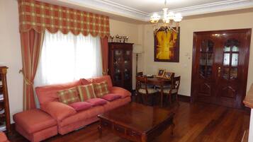 Una sala de estar con un sofá, una mesa de centro de madera, una mesa de comedor con sillas, una araña y una puerta con paneles de vidrio.