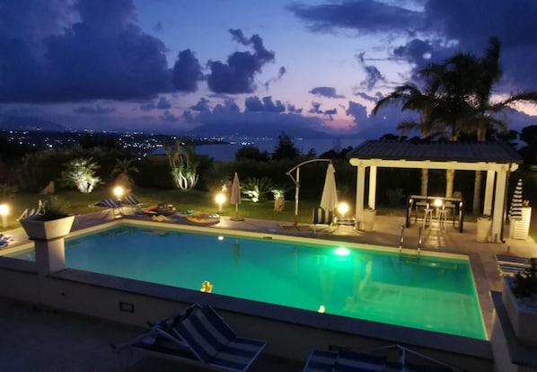 A swimming pool with lounge chairs, a covered seating area, and a view of the city at dusk.