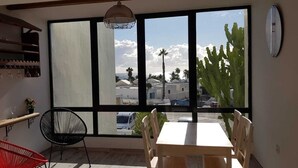A room with a large window, a wooden table, a red chair, a black chair, and a hanging light fixture. - Residence Playa Roca Costa Teguise "LANZAROTE" (Costa Teguise)