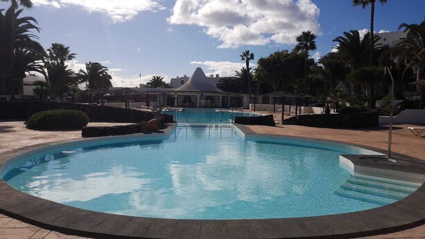 A large, clear swimming pool with a central island and steps leading into the water, surrounded by palm trees and a building with a dome-shaped roof in the background. - Residence Playa Roca Costa Teguise "LANZAROTE" (Costa Teguise)