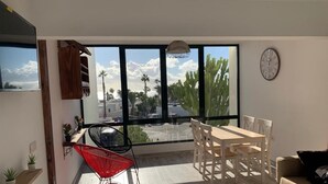 A modern living room with a large window, a dining table, a red chair, and a sofa. - Residence Playa Roca Costa Teguise "LANZAROTE" (Costa Teguise)
