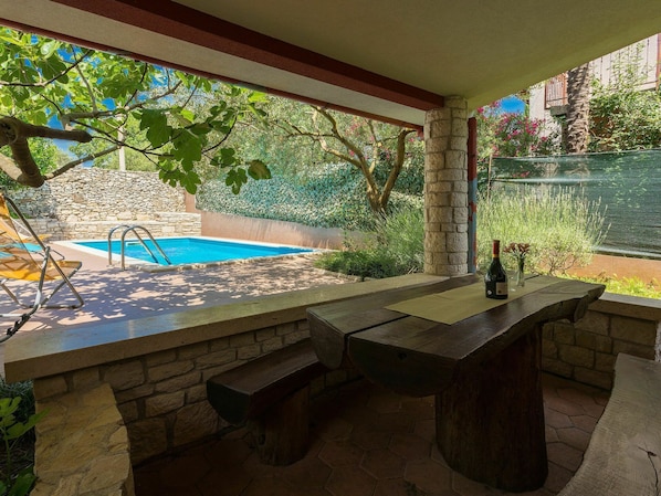 An outdoor pool area with a stone bench and a table, surrounded by greenery and a stone wall.