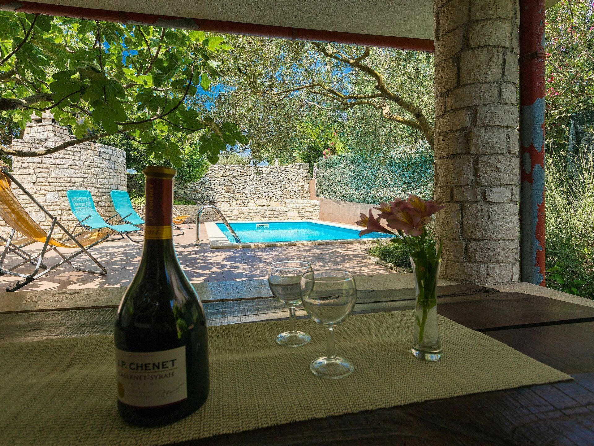 Eine Weinflasche, zwei Gläser und eine Vase mit Blumen auf einem Tisch mit Blick auf einen Swimmingpool und Liegestühle.
