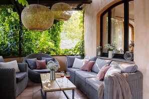A cozy outdoor seating area with wicker furniture, a table with wine glasses, and a view of greenery through a window.