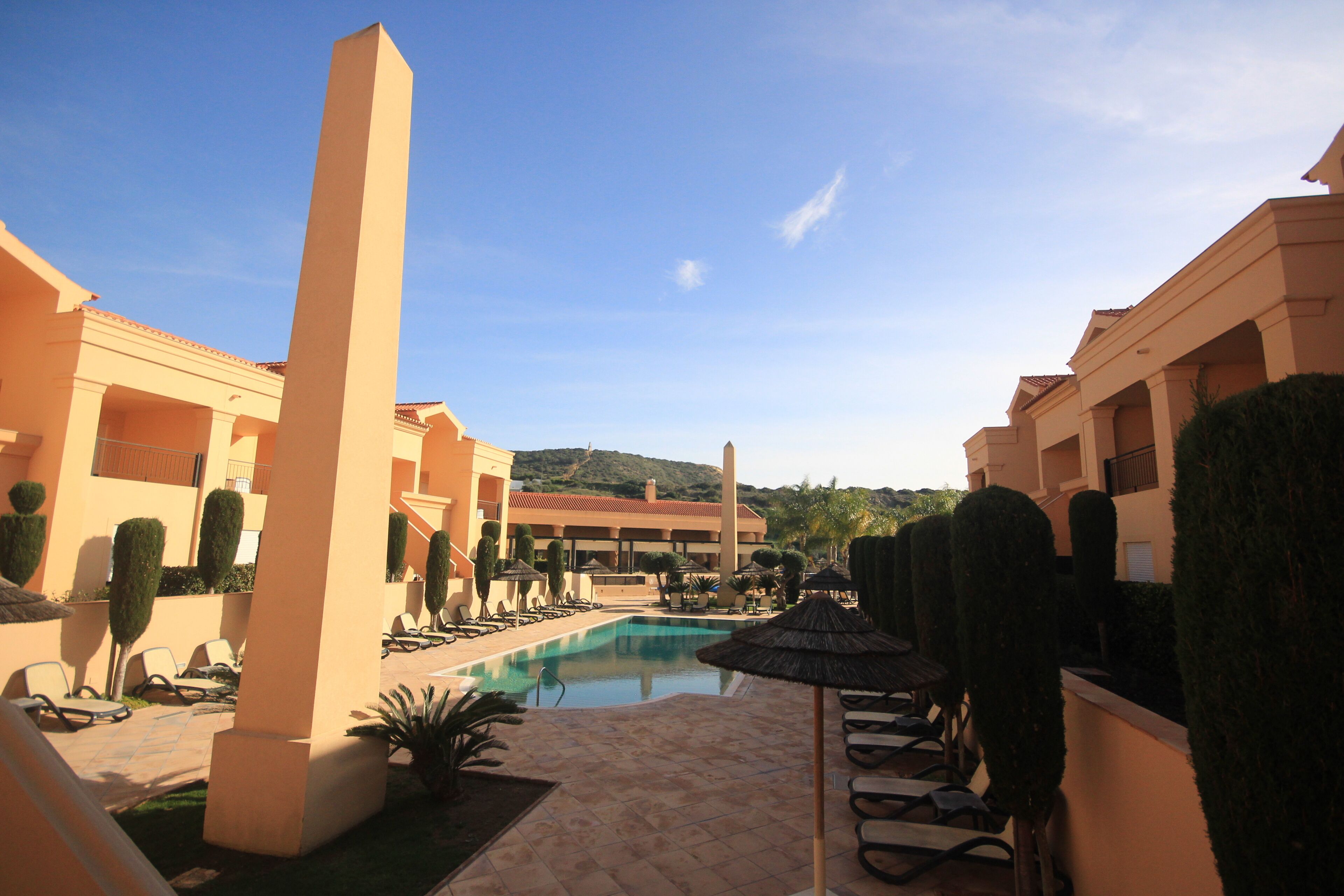 A pool area with lounge chairs and umbrellas, surrounded by buildings with a Mediterranean architectural style.
