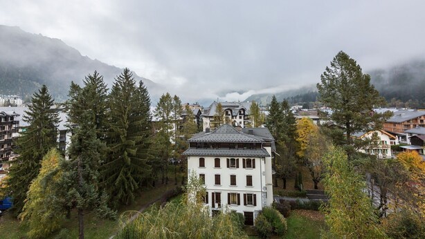 Un edificio blanco con techo inclinado, rodeado de árboles y con montañas al fondo.