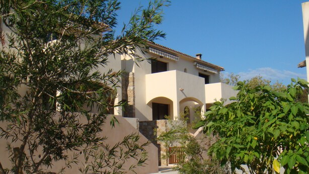 A building with a beige facade, stone accents, and a tiled roof. There are arched doorways and a balcony with a railing.