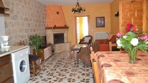 A cozy room with a stone-patterned floor, a fireplace, a dining area with a table and chairs, and a bed with a floral bedspread. - Bocanegra Rural Cottage for 10 people (Canicosa de la Sierra)