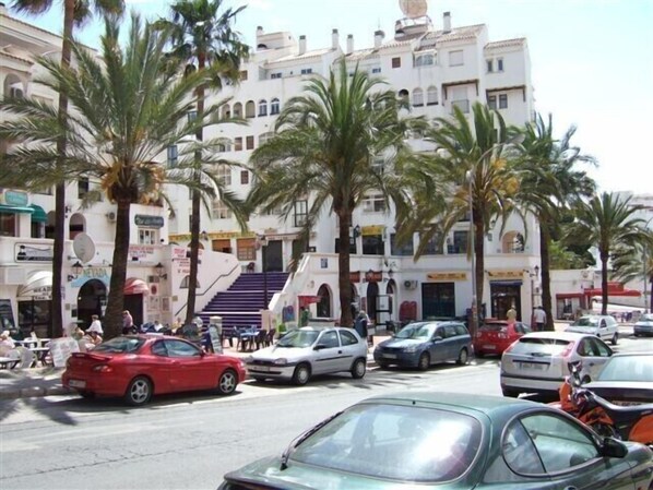 A street scene with parked cars, palm trees, and a building with multiple floors and balconies.
