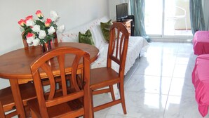 A dining area with a wooden table and chairs, a sofa with a floral cushion, and a TV on a stand. - A beautiful studio apt overlooking pools ' Minerva.Jupiter @ Bonanza Square ' (Benalmadena)
