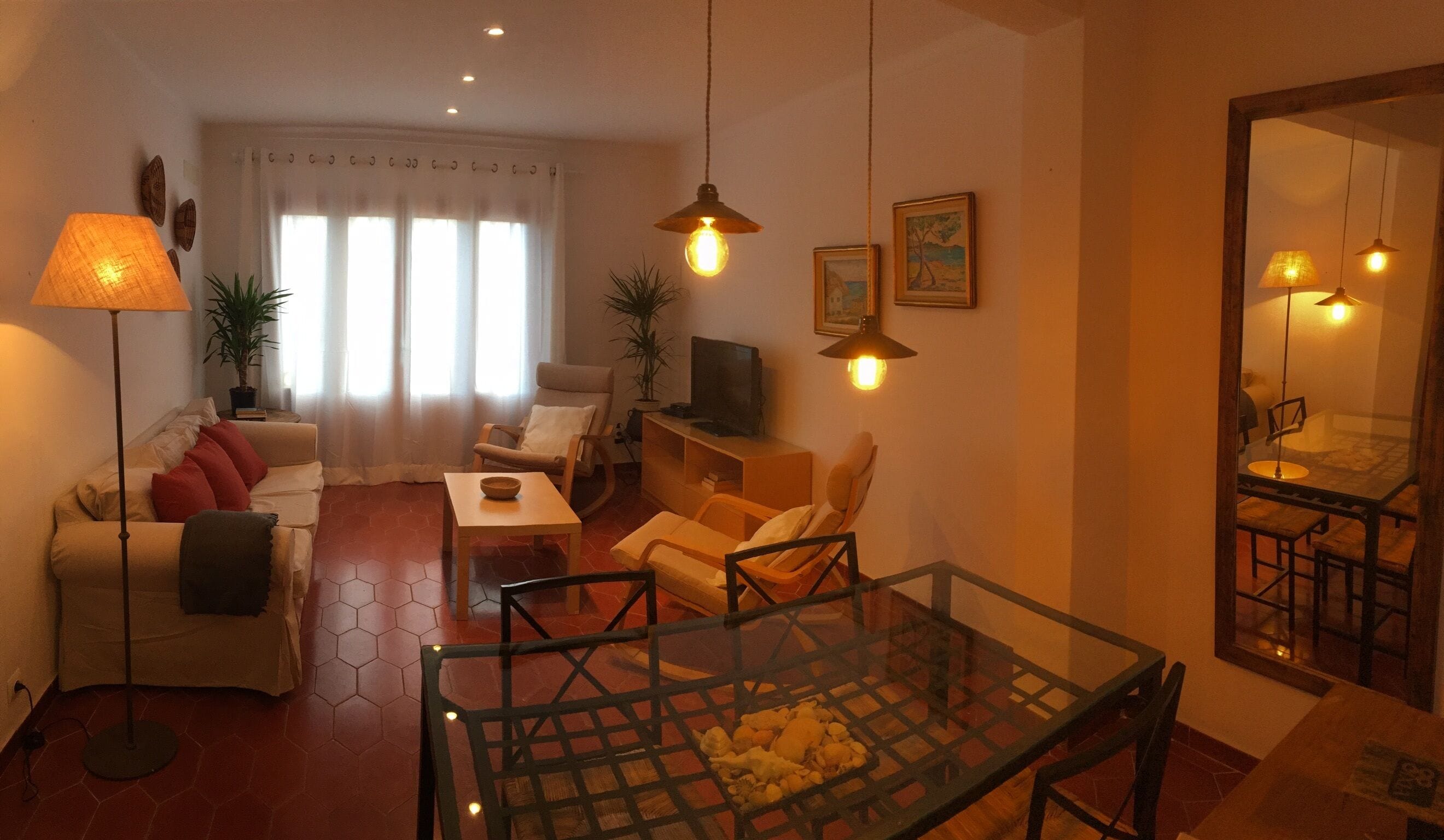 A living room with a sofa, armchairs, a glass coffee table, and a TV. There are plants, a lamp, and a mirror on the walls.