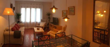 A living room with a sofa, armchairs, a glass coffee table, and a TV. There are plants, a lamp, and a mirror on the walls.