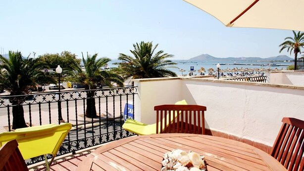 Eine Terrasse mit einem Holztisch, gelben Stühlen und einem großen Sonnenschirm. Die Terrasse bietet Blick auf einen Strand mit Palmen und eine Aussicht auf das Meer und die entfernten Berge.