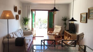 Ein Wohnzimmer mit einer Couch, Sesseln, einem Glastisch und einem TV-Schrank. Üchtisgefäße und eine Hängeleuchte sind ebenfalls vorhanden.